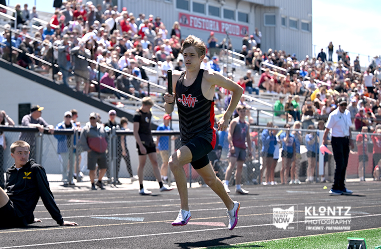 Male sprinter in black and red uniform runs on a track while holding a baton during a relay race, with a crowded stadium in the background.