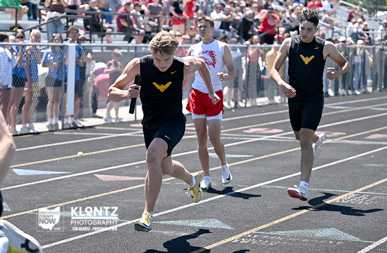 Three male track sprinters race on an outdoor track; the lead runner in black with a yellow logo carries a baton as others chase him with spectators behind the fence.