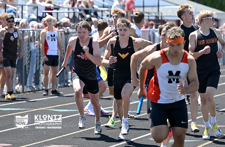 Track relay race: sprinters sprint with batons,crowd behind fencing at a sunny stadium.