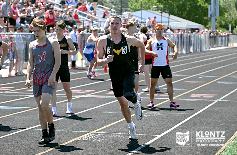 Group of male sprinters racing on an outdoor track; lead runner in black with a knee brace and bib number 3, others behind, with spectators in the stands.