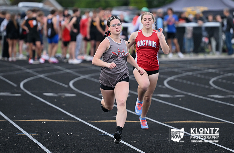 Two female sprinters race on a track with a cheering crowd in the background, lead runner in gray top.urrent behind in red Bucyrus uniform.