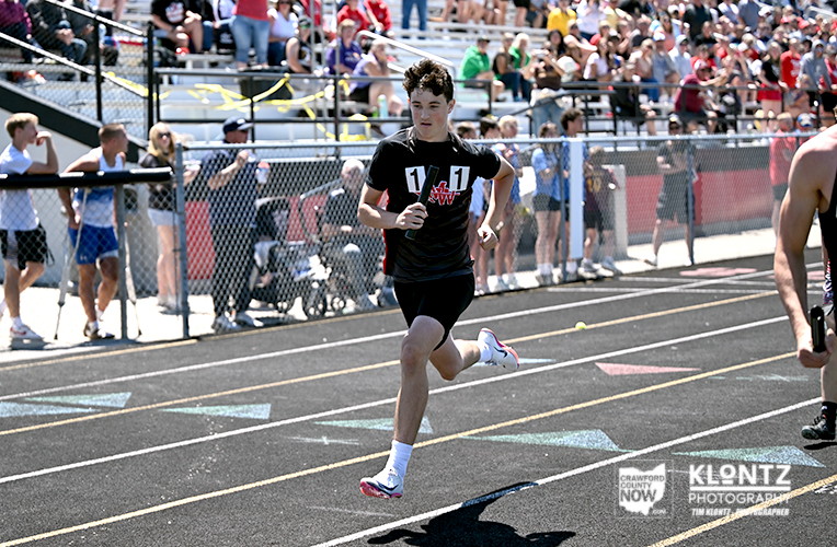 Young male sprinter in a black shirt and white socks runs on an outdoor track as a crowd watches from behind a chain-link fence.
