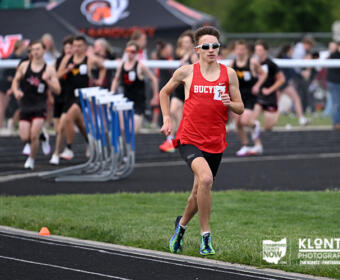 Male sprinter in a red Bucyrus jersey leads a race on an outdoor track, with teammates and spectators blurred in the background.