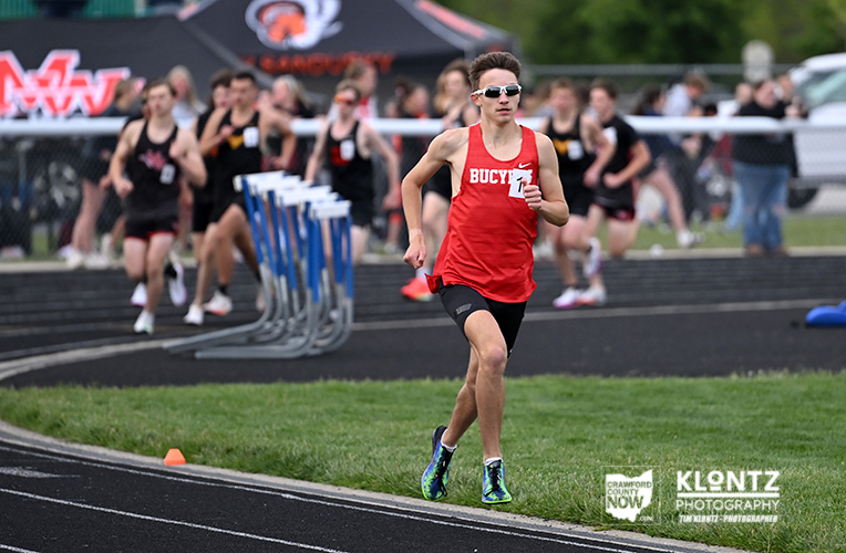 Male sprinter in a red Bucyrus jersey leads a race on an outdoor track, with teammates and spectators blurred in the background.