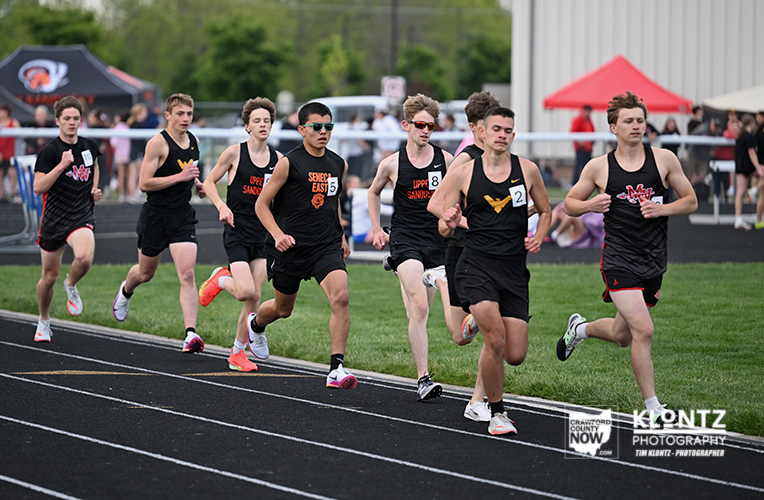 Male track athletes sprint from the starting line on a black oval track; runners wear black uniforms with orange logos, with spectators and tents in the background