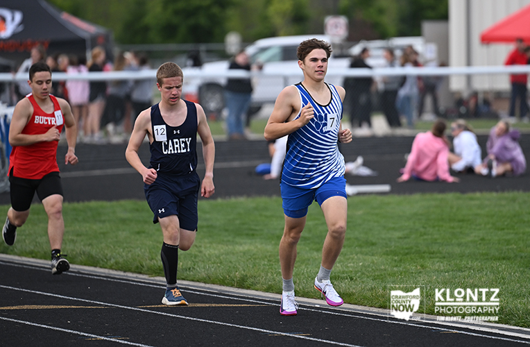 Three male sprinters race on an outdoor track; front runner in a blue-and-white striped uniform leads, others behind him.