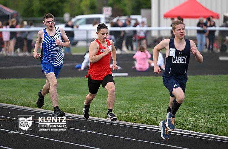 Three male sprinters race on an outdoor track during a meet; red jersey center, blue uniforms left and right, spectators in the background.