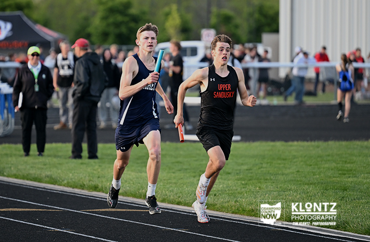 Two male relay runners race side by side on a track, each holding a baton as they sprint toward the finish line.