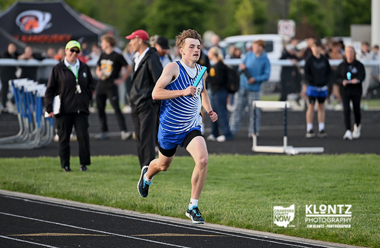 Young male sprinter in a blue striped uniform races on an outdoor track, spectators and event tents in the background.