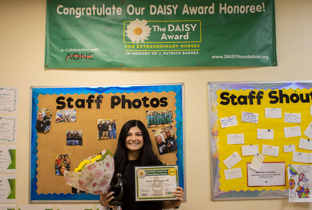 Smiling nurse holds a bouquet and DAISY Award certificate in front of a staff photos board and a congratulatory banner.