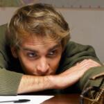 Young man with blond hair rests his arms on a desk, gazing intently at a small wooden sculpture on the right.