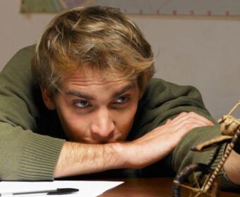 Young man with blond hair rests his arms on a desk, gazing intently at a small wooden sculpture on the right.