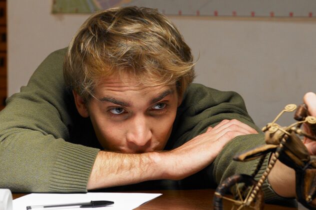 Young man with blond hair rests his arms on a desk, gazing intently at a small wooden sculpture on the right.