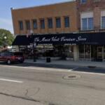 Front of The Amish Vault Furniture Store on a sunny street with a black awning, white lettering, and parked cars nearby.