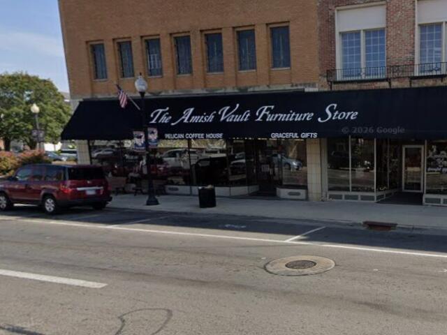 Front of The Amish Vault Furniture Store on a sunny street with a black awning, white lettering, and parked cars nearby.