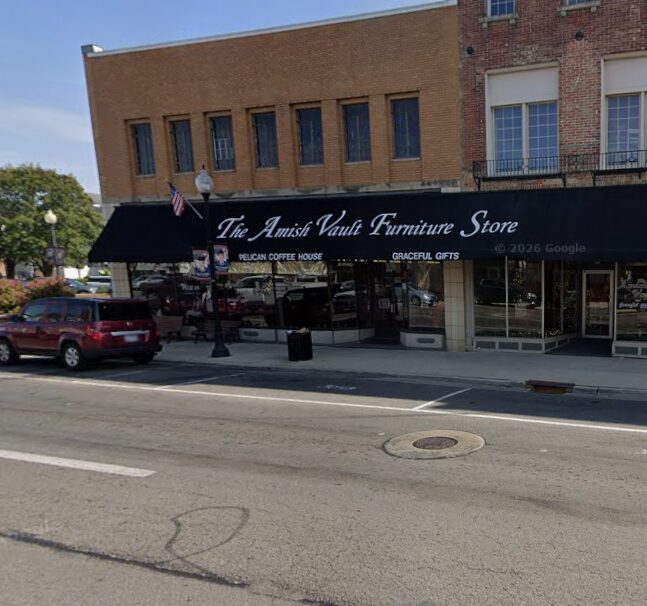 Front of The Amish Vault Furniture Store on a sunny street with a black awning, white lettering, and parked cars nearby.