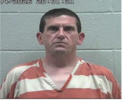 Mugshot of an adult man in an orange-and-white prison jumpsuit facing the camera against a gray wall.