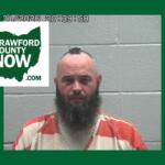 Mugshot of a bearded man in an orange prison jumpsuit against a gray cinder-block wall, with a Crawford County Now logo overlay on the left.