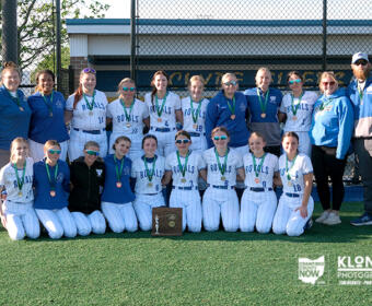 Girls softball team in white Royals uniforms posing with medals and a trophy on a baseball field.