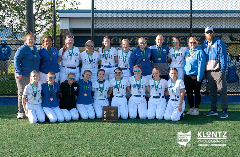 Girls softball team in white Royals uniforms posing with medals and a trophy on a baseball field.