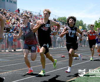 Group of young male sprinters racing on a track with a crowded stadium in the background, spectators cheering.