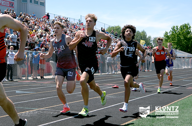 Group of young male sprinters racing on a track with a crowded stadium in the background, spectators cheering.