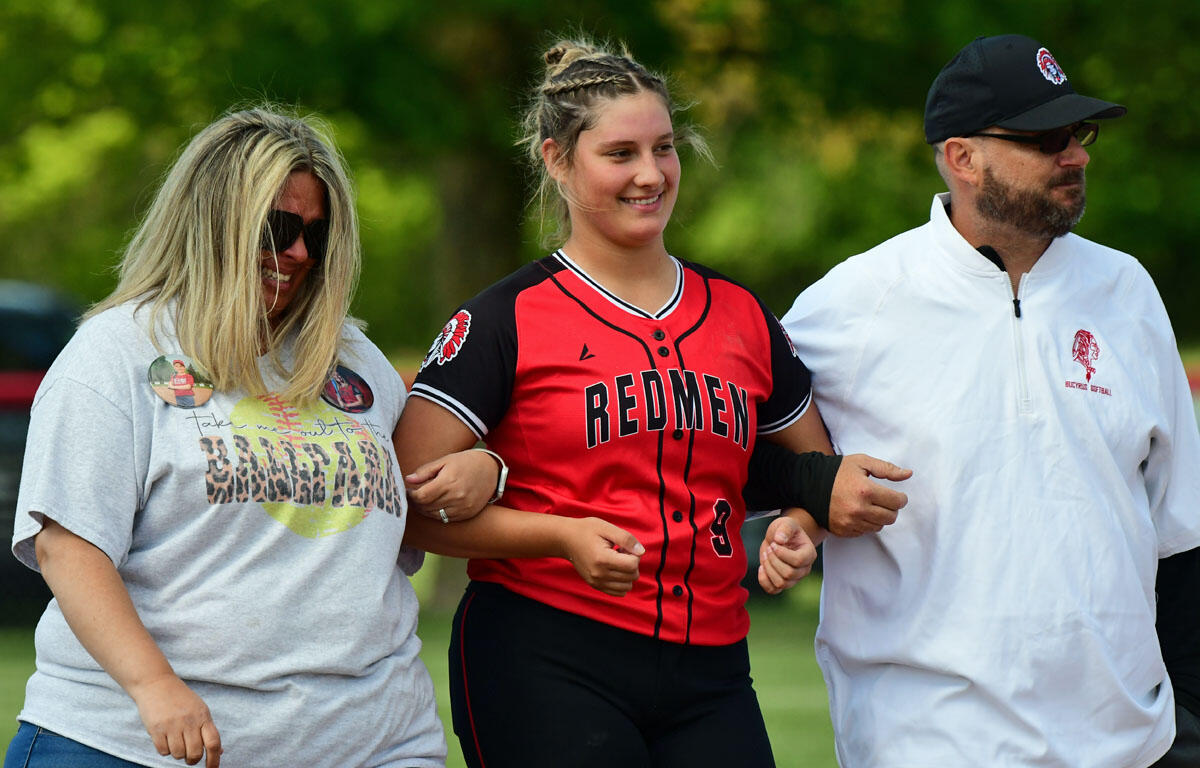 Softball player in a red and black 'Redmen' jersey walks arm-in-arm with two adults outdoors on a sunny field.