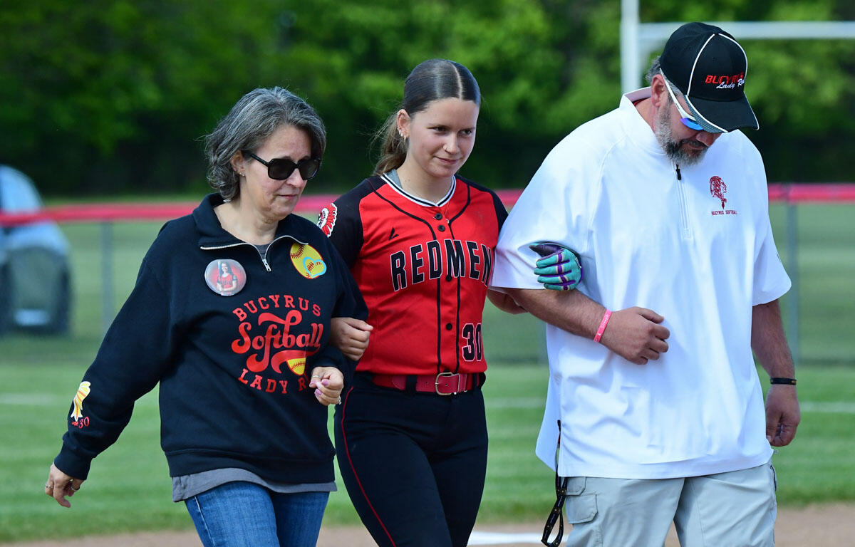 Softball player in red and black uniform being guided off the field by two adults.
