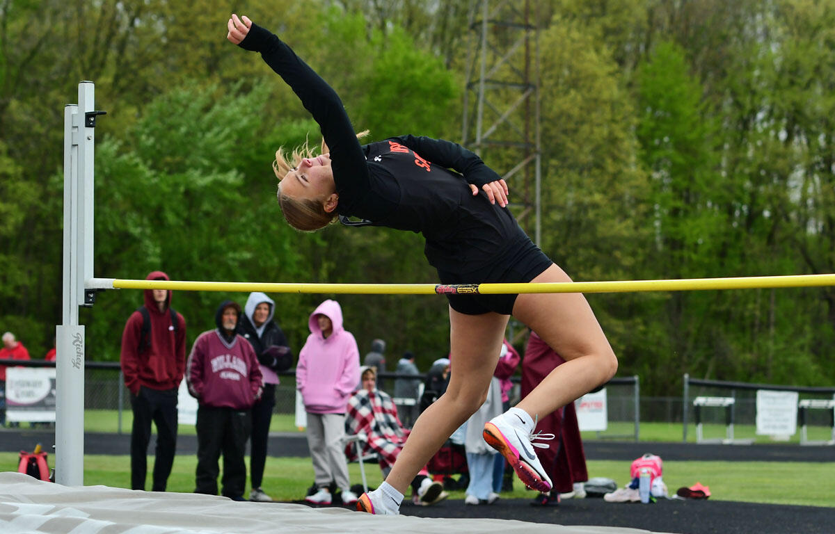 Female high jumper in a black outfit clears a yellow bar over a mat on an outdoor track, spectators watching in the background.