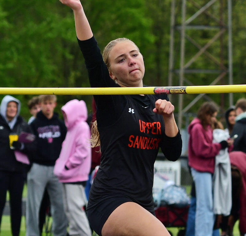 Female athlete clears a yellow high jump bar during a track meet, lifting one arm as she vaults over.