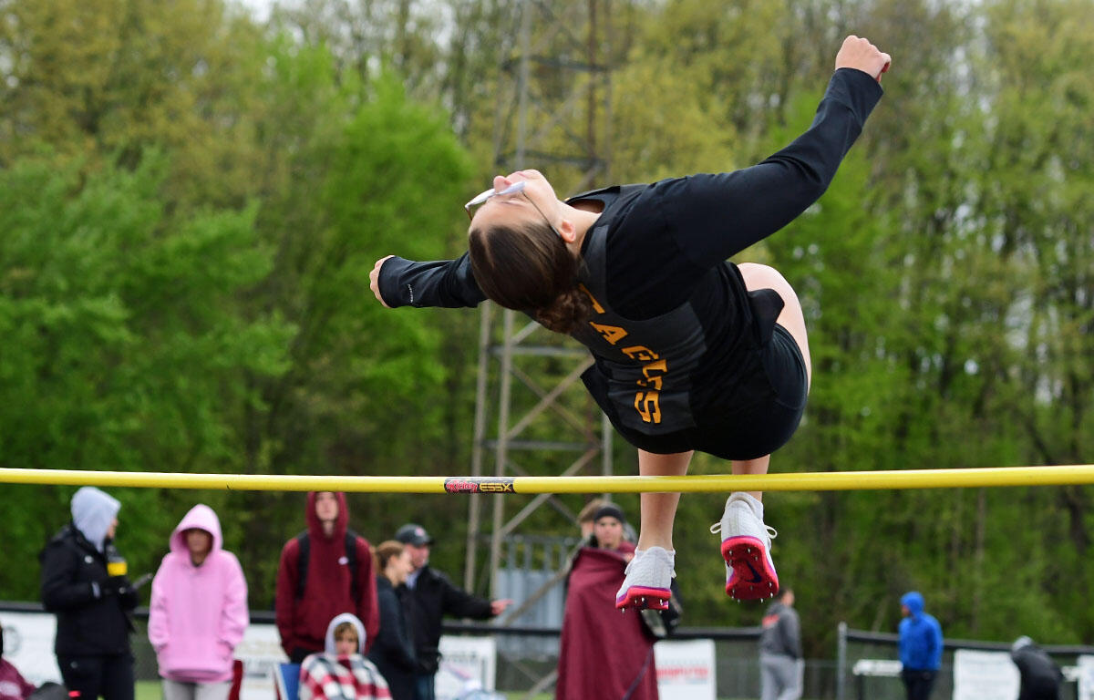 Athlete clears a yellow high jump bar during a track and field meet, spectators watching in the background.