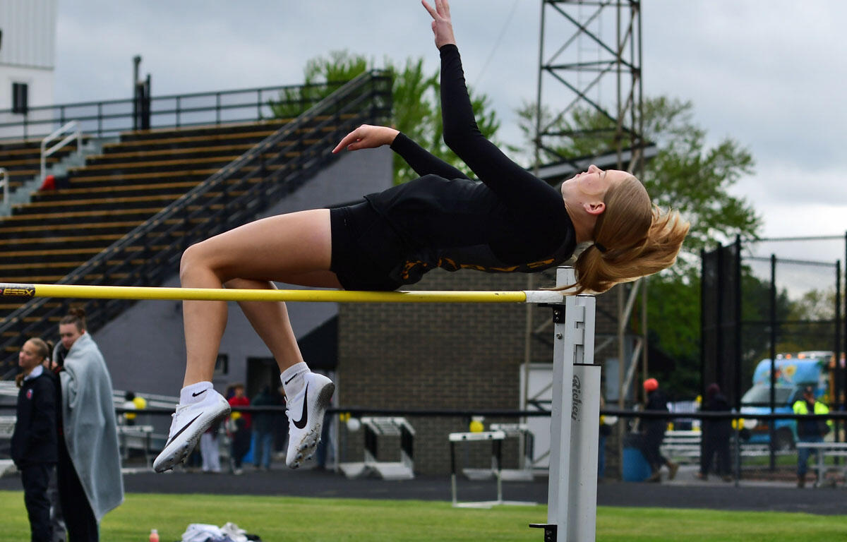 Female athlete in black leotard clears a high jump bar in a stadium, mid-air with back arched and arms extended.
