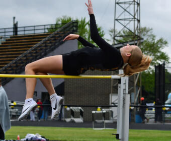 Female athlete in black leotard clears a high jump bar in a stadium, mid-air with back arched and arms extended.