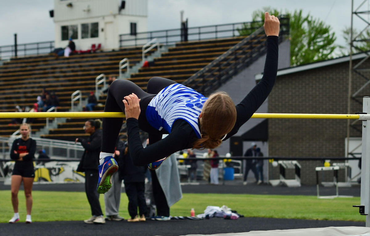 Female athlete clears a high jump bar in a blue-and-white top and black leggings at an outdoor track stadium, mid-air over the yellow bar.