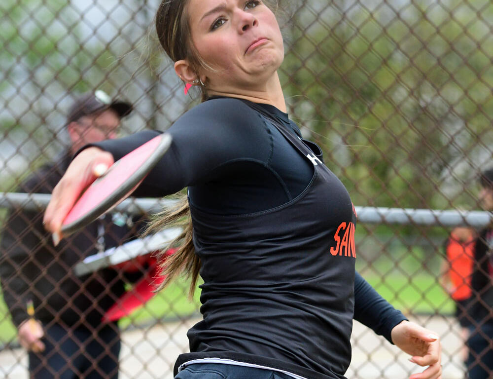 Female athlete in a black long-sleeve jersey throwing a pink frisbee on a field behind a chain-link fence; focused expression shows effort.