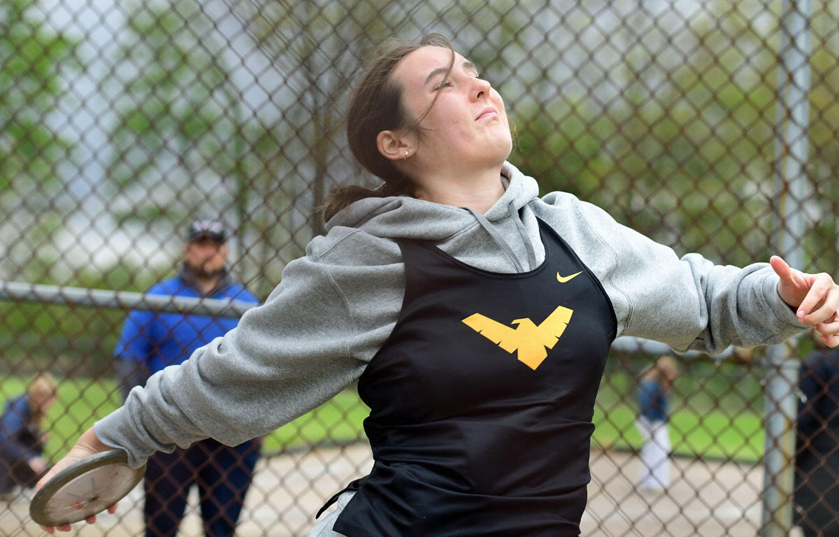 Athlete in gray hoodie and black singlet throws a frisbee during a game behind a chain-link fence, wind in her hair and focused expression.
