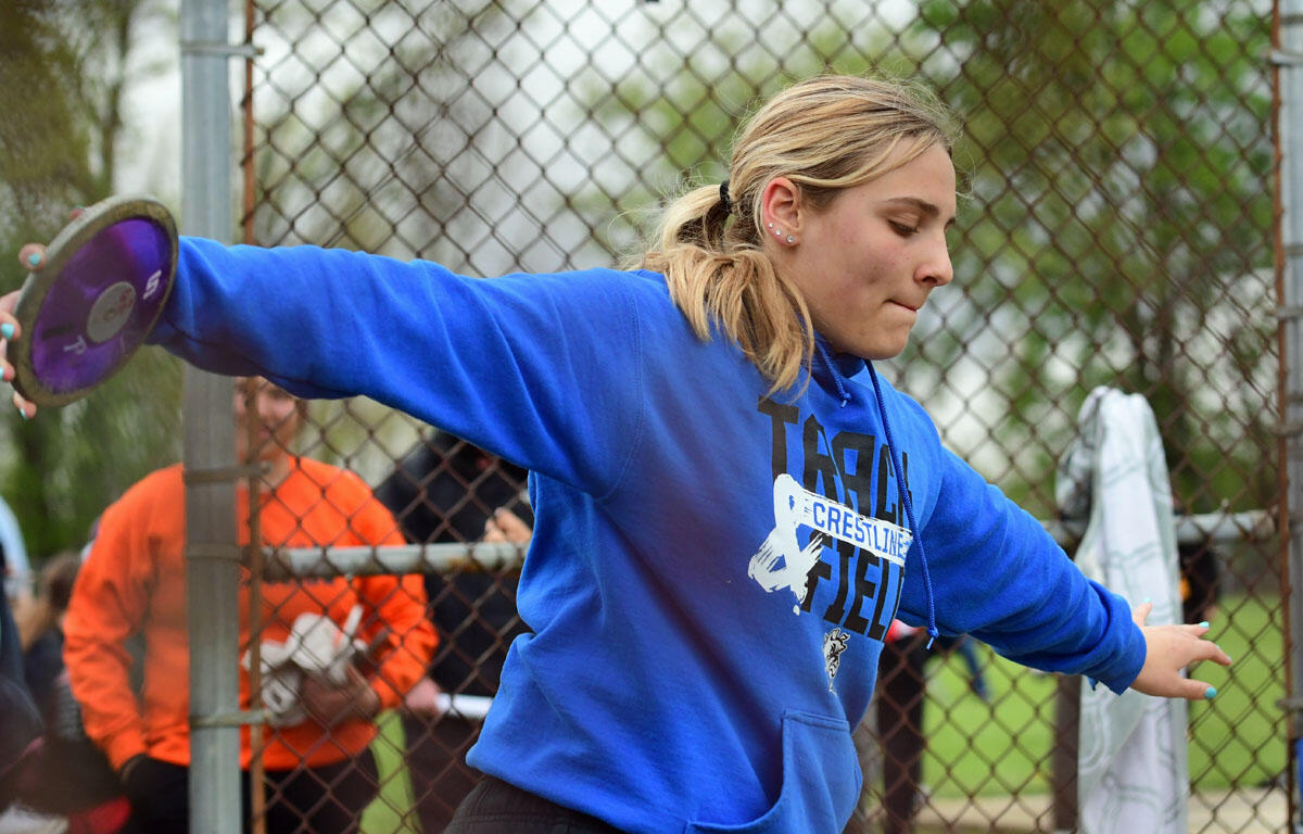 Young woman in a blue hoodie throwing a discus at an outdoor track field, with a chain-link fence behind.