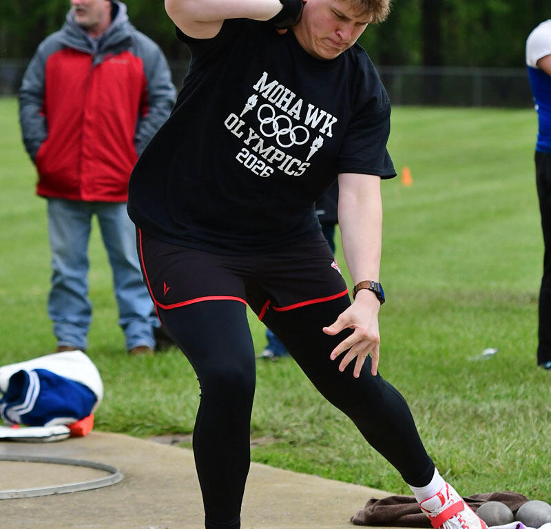 Male athlete in a black shirt throwing on a track/field circle, spectators in the background, outdoors on a grassy field.