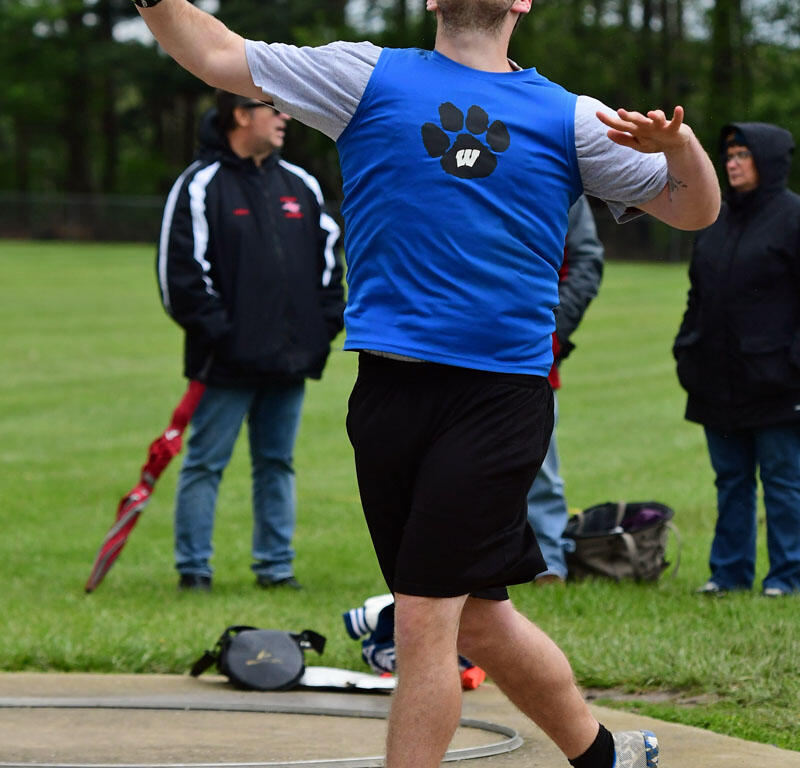 Male athlete in a blue shirt throws a shot put from the throwing circle as spectators watch on a grassy field.