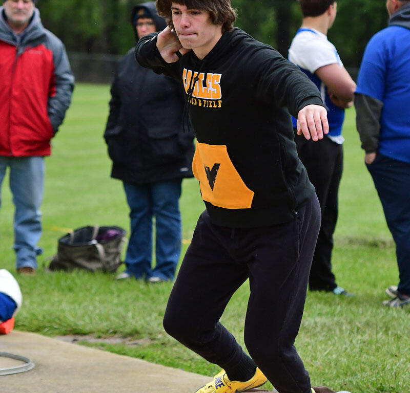 Young male athlete in a black hoodie and yellow Nike shoes is winding up for a throw on a track-and-field throwing circle, spectators in the background trustees the field.