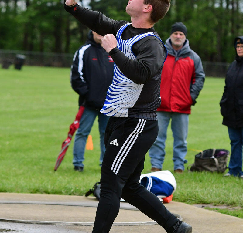 Male hammer thrower in a blue striped singlet releasing the hammer on the throwing circle, with onlookers in the background on a grassy field.