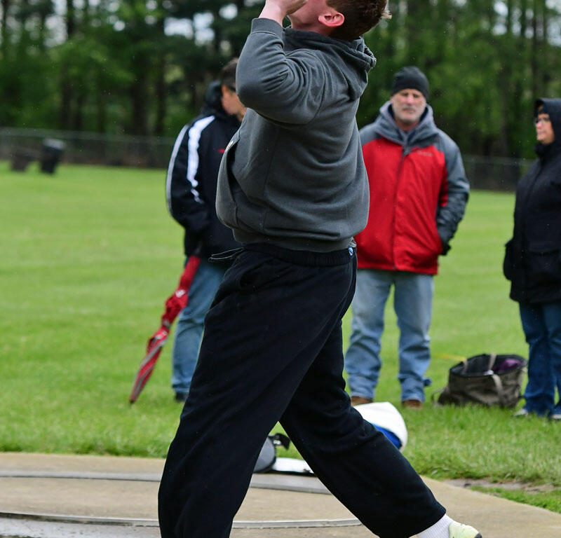 Man in a gray hoodie throws a shot put during a track and field event on a grassy field, with spectators in the background.