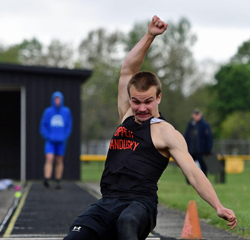 Male track athlete in a black sleeveless shirt lands in the sand pit during a long jump attempt.