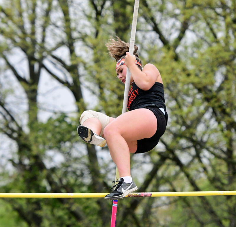 Female pole vaulter in mid‑flight, gripping a tall pole as she clears a yellow bar outdoors with trees in the background.
