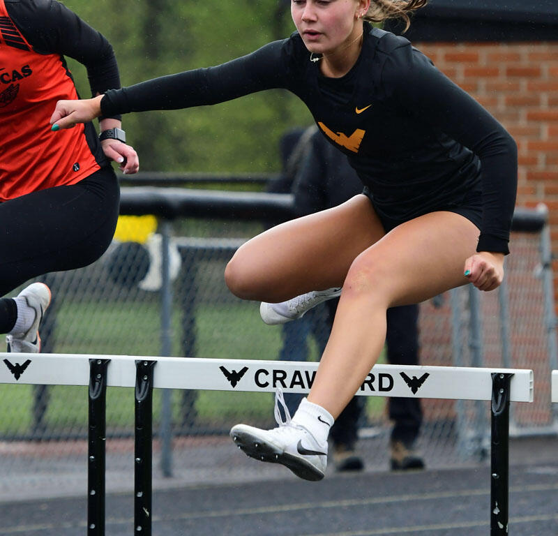 Female track athlete in black kit clears a hurdle mid-race on a track field with a competitor in red nearby