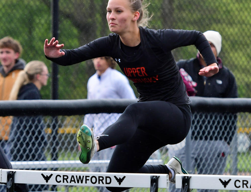 Female hurdler in black athletic wear clears a hurdle on a track, with spectators behind a chain-link fence.