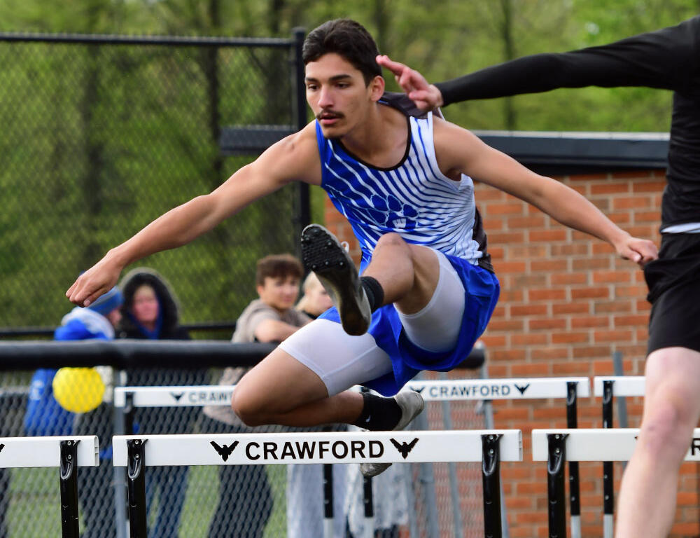 Male sprinter in blue-and-white uniform clears a track hurdle mid-air, hurdling over a Crawford barrier with spectators in the background and greenery beyond.