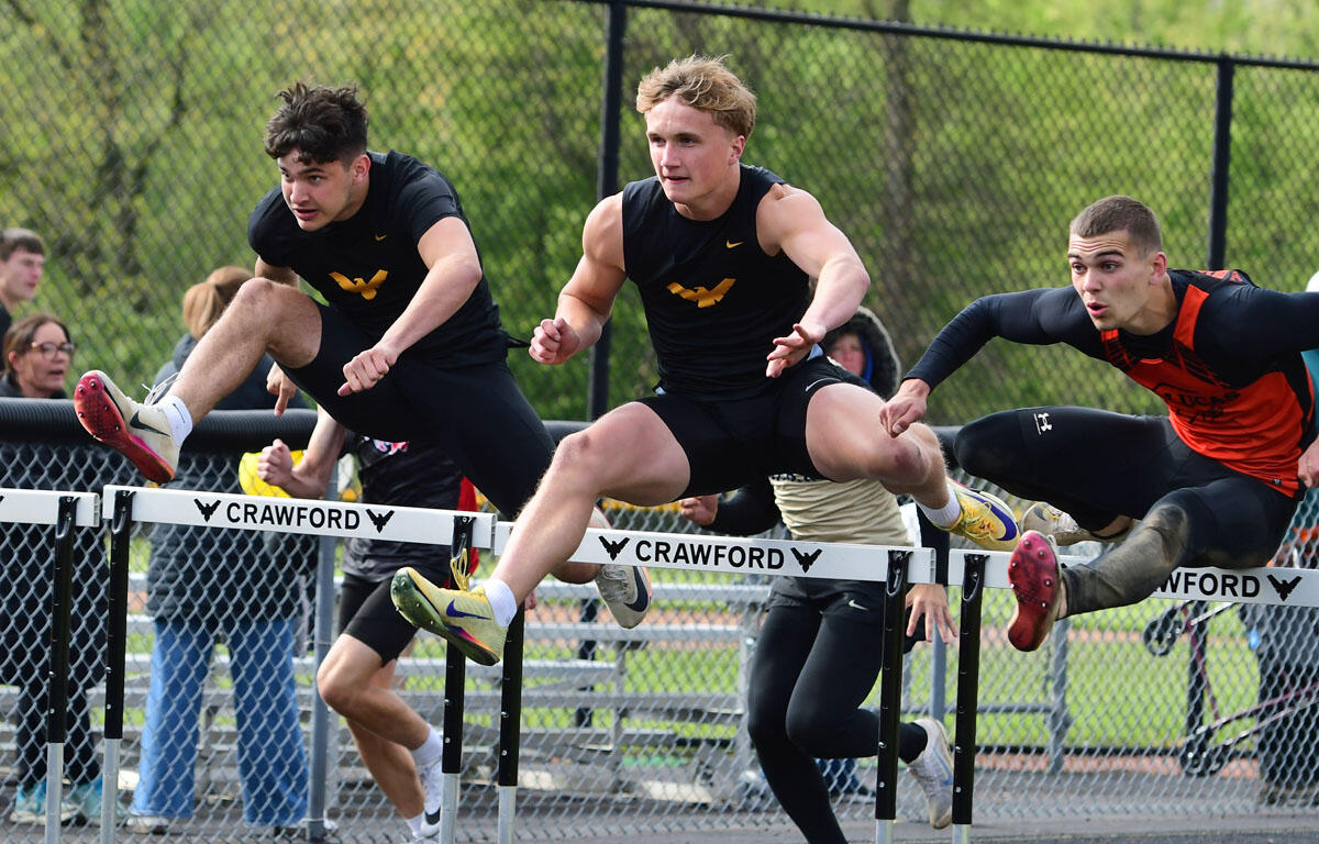 Three male hurdlers leap over a track hurdle during a race, wearing black and orange uniforms, with a chain-link fence and spectators in the background.