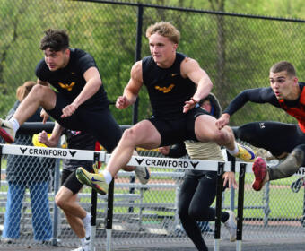 Three male hurdlers leap over a track hurdle during a race, wearing black and orange uniforms, with a chain-link fence and spectators in the background.