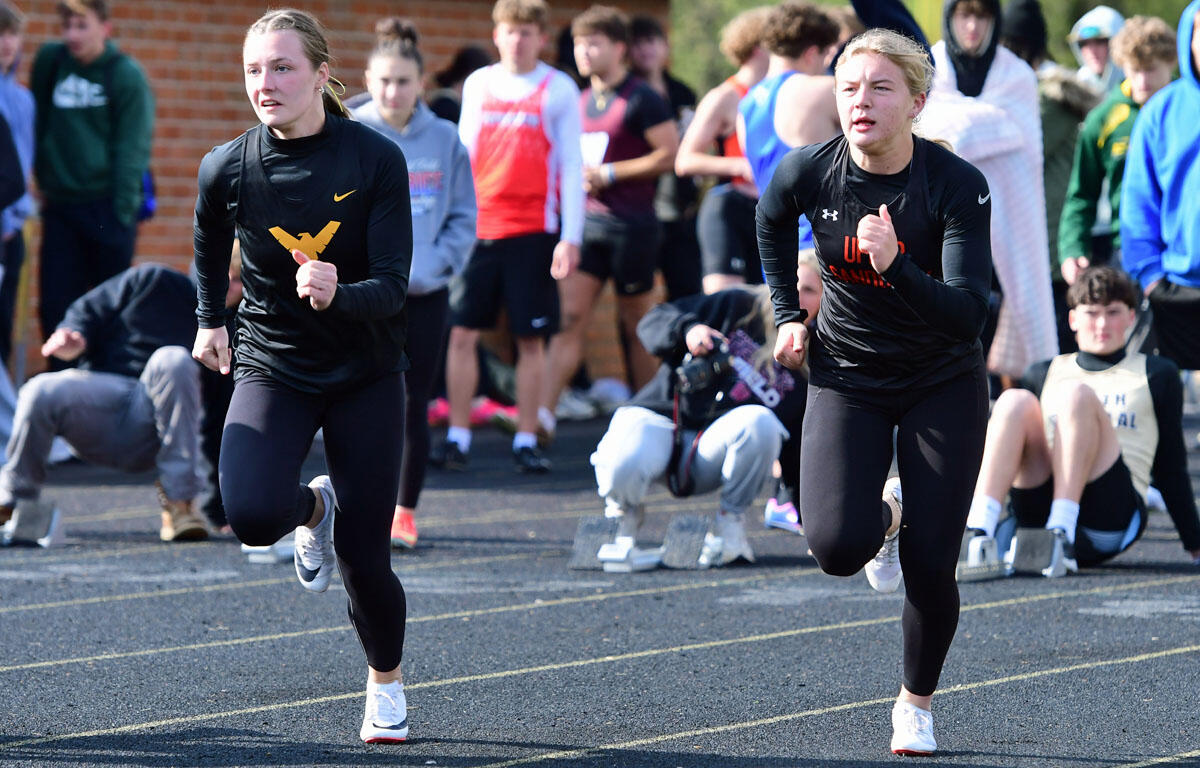 Two young women in black athletic outfits sprint on a track as a crowd watches in the background.